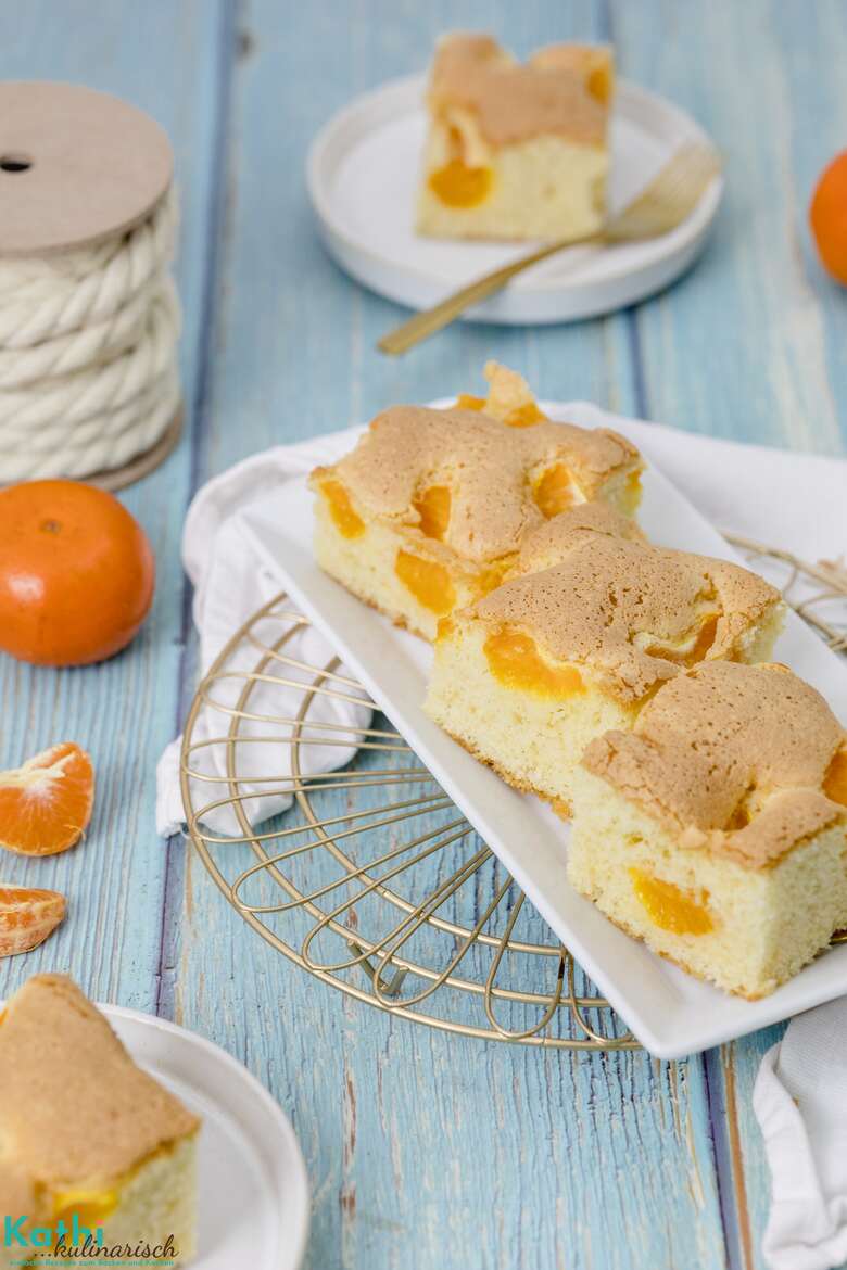 Auf einem blauen Holztisch wurde gerade auf einer weißen Platte ein saftiger Buttermilchkuchen mit Mandarinen serviert. Die goldenen Gabeln liegen schon bereit und zwei Früchte sind auch zu sehen.